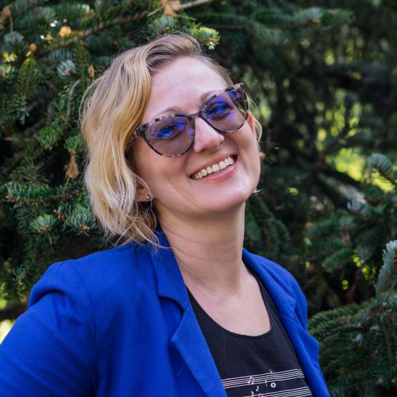Jennifer Johnson in a blue blazer with a black shirt underneath smiling at the camera infront of a pine tree